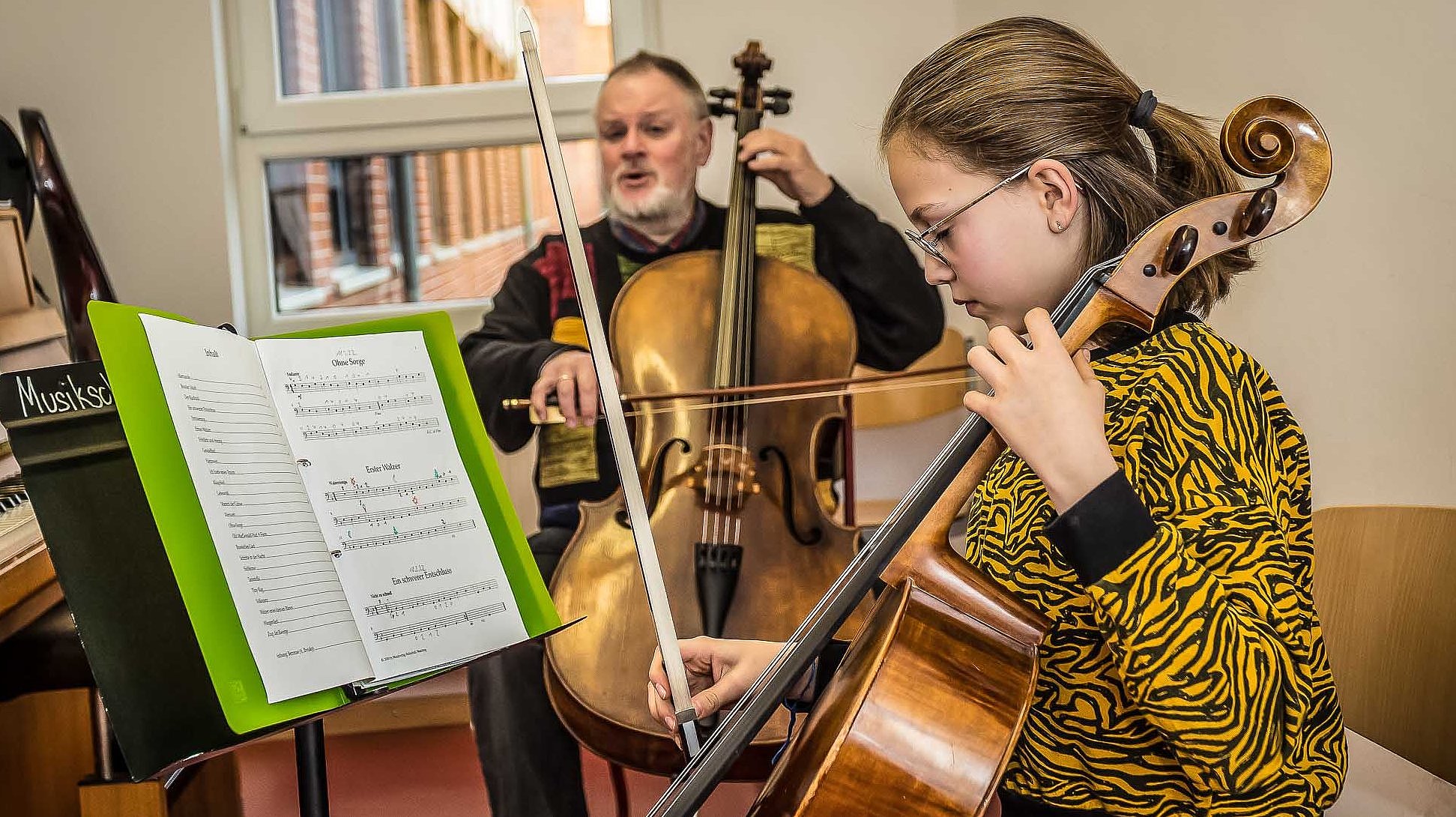 Jugendliche spielt konzentriert Cello vor einem Notenständer, im Hintergrund der Cello spielende Lehrer